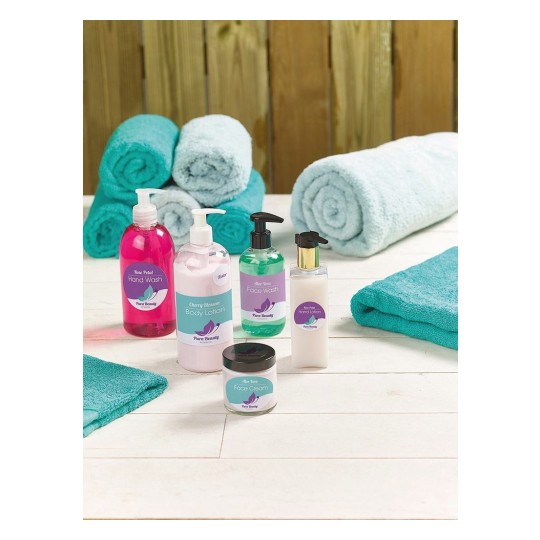 The image displays a collection of beauty and skincare products including hand wash, body lotion, face wash, and face cream, arranged beautifully on a white wooden surface with teal towels in the background.