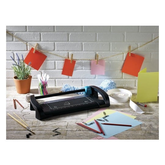 The image features a crafting workspace with a paper trimmer, colorful papers, clips, and stationery tools. There are red, pink, and yellow paper sheets displayed along a string, with a potted plant and various stationery items scattered on a wooden table.