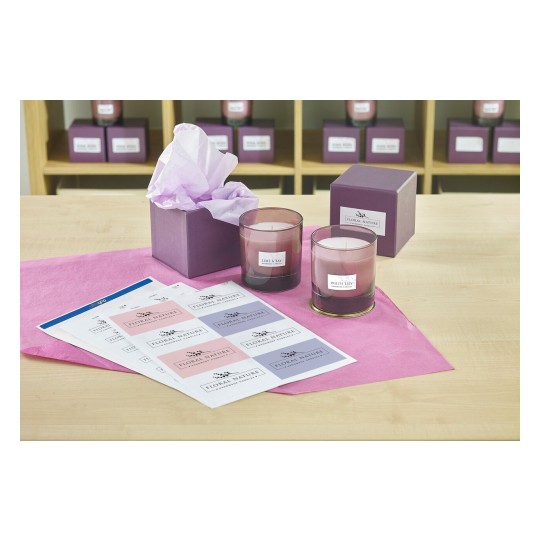 The image features two scented candles labeled 'Lime & Bay' and 'White Lily' on a table. There is a purple gift box, pink tissue paper, and sheets with various candle labels. The background shows shelves with more candles.