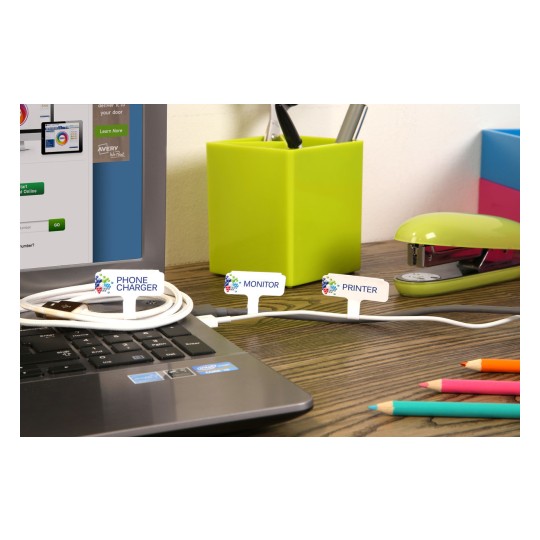 The image shows a laptop on a wooden desk with labeled cords for a phone charger, monitor, and printer. There are colorful pencils, a green stapler, and a green container with pens in the background.