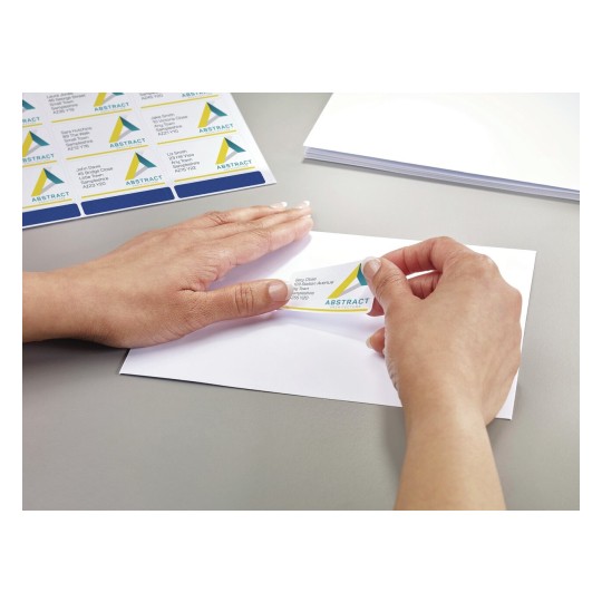 The image shows a person placing a label on an envelope. The label has a design with the word 'ABSTRACT' and includes a name and address. There are additional labels visible on a sheet, and blank envelopes are on the table.