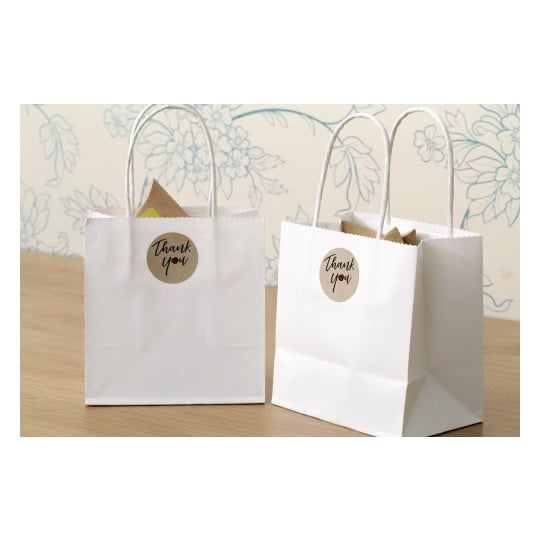 The image features two white gift bags with 'Thank You' labels on them. They are placed on a wooden surface, with a floral-patterned wall in the background. The bags contain items that are not fully visible.