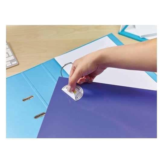 An individual is placing a circular sticker on a purple folder, with additional blue folders and a keyboard visible in the background.