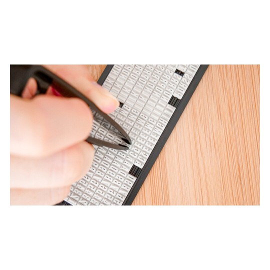 The image shows a hand using tweezers to pick up letters from a flat surface containing many small plastic letter tiles arranged in rows.