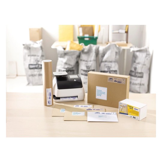 The image shows a packaging station with a printer, rolled paper, various envelopes, a cardboard box, and franking labels. In the background, there are bags, likely for shipping materials.