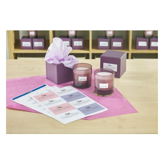 The image shows two scented candles placed in front of a purple gift box with tissue paper. There are also labels laid out on the table, indicating different candle scents. The background contains more purple boxes.