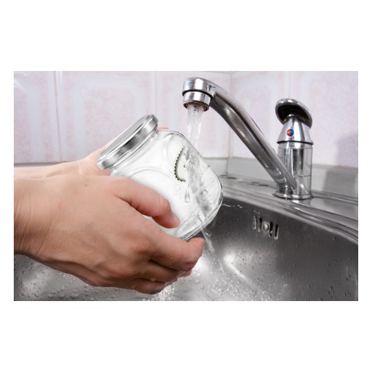 The image shows a hand holding a glass jar under running water from a kitchen faucet, while water splashes into the sink.