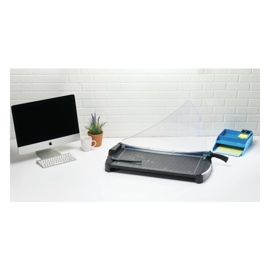 The image shows a workspace featuring an iMac computer, a plant in a pot, a white mug, a paper cutter, and a blue organizer with stationery items on a white table against a white brick background.