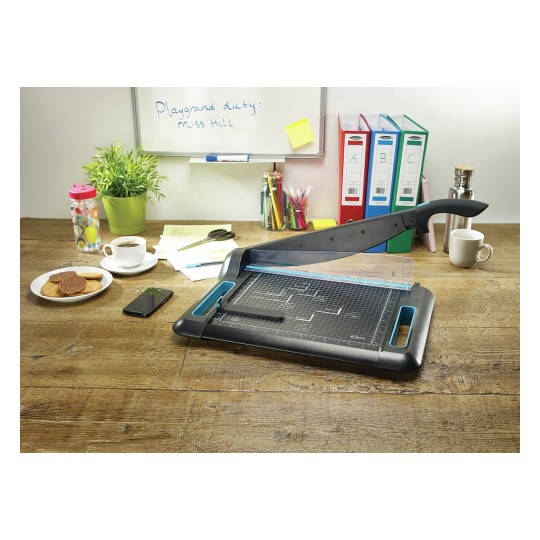 The image shows an office workspace with a paper cutter, a cup of coffee, cookies on a plate, a smartphone, colorful pens, and a potted plant, with notes and file organizers in the background.