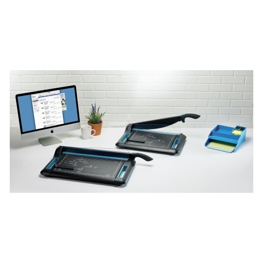The image shows a workspace with two paper trimmers, a computer displaying a webpage, a potted plant, a white mug, and a blue organizer with stationery items.