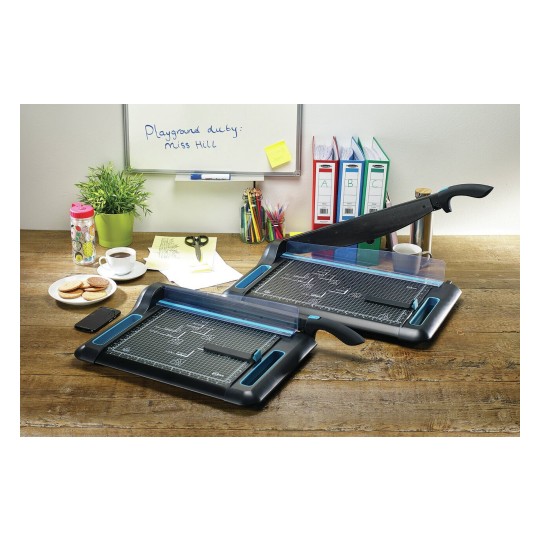 The image shows a workspace with two paper trimmers, a cup of coffee, a plate of cookies, scissors, colorful pens, a plant, and a whiteboard with a note.