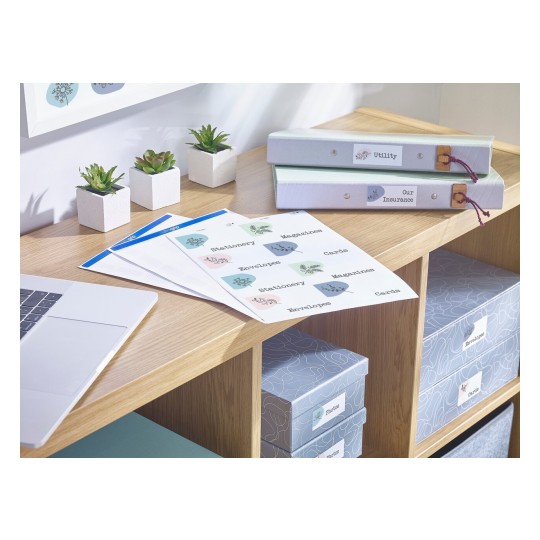 The image showcases a workspace featuring potted plants, files in green and blue binders labeled 'Utility' and 'Our Insurance', printed sheets with various labels, and storage boxes for photos and cards on a wooden table.