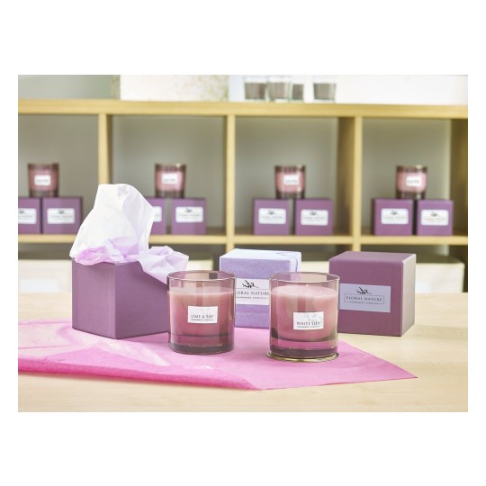 The image features various scented candles displayed on a table, with two prominent candles in glass holders labeled 'Lime & Bay' and 'White Lily' surrounded by gift boxes in shades of purple. The background shows additional candles organized on a shelf.