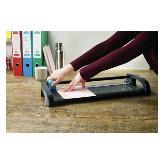 A person cutting a pink sheet of paper using a paper trimmer on a wooden desk. There are binders, a cup, and a stainless steel water bottle in the background.