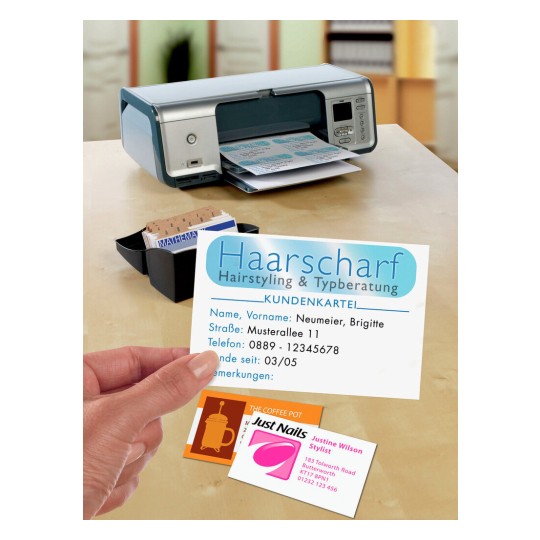 The image shows a printer on a tabletop with business cards and a customer card for a hairstyling service. The customer's card displays the name Neumeier, Brigitte, with contact information. Other cards on the table are for Just Nails and The Coffee Pot.