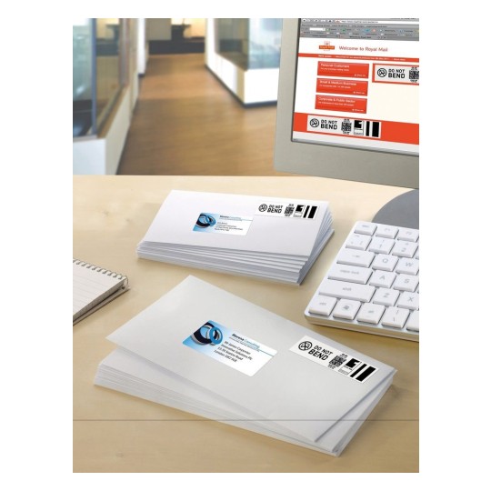 The image shows a stack of white envelopes with labels on them, placed on a desk with a keyboard and a notepad. A computer screen displays the Royal Mail website, indicating services for personal and business mail.