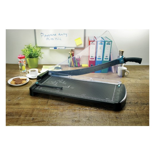 The image shows a paper cutter on a wooden table. There's a plate of cookies, a cup of coffee, a plant, and some stationery including binders and pens in the background. A whiteboard with a note is also visible.