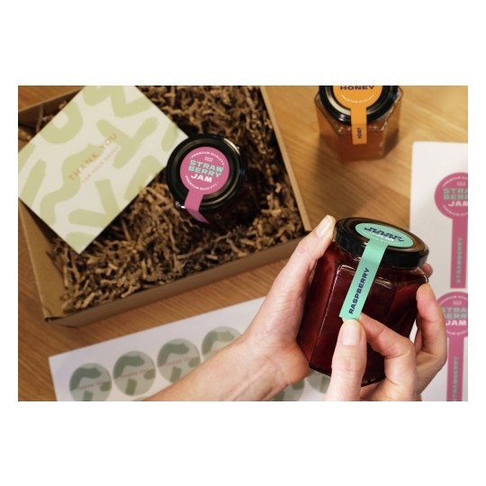 The image shows a gift box containing jars of raspberry jam and strawberry jam, with a thank you card and stickers. A hand is holding a jar that has a green label indicating it's raspberry.