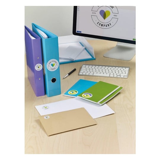 The image features an organized workspace with colorful binders, notebooks, and envelopes on a wooden desk. A computer monitor displays a logo design for a company. Items include blue, green, and purple binders, several envelopes, and a keyboard.
