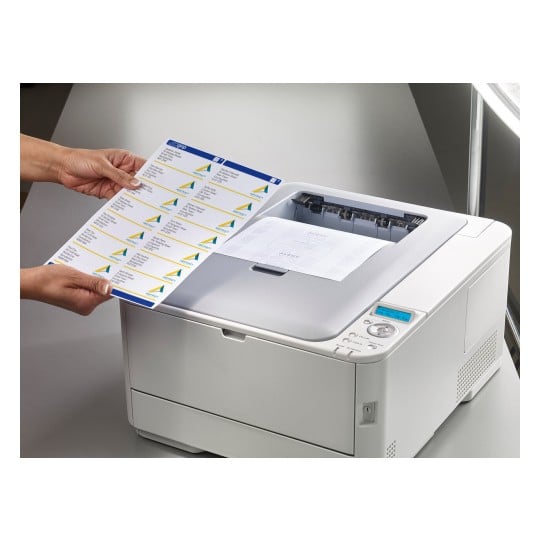 The image shows a person holding a sheet of printed labels above a printer, which has a document partially printed inside. The printer is modern and on a desk.