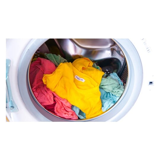The image shows a washing machine full of clothes in various colors, including a yellow t-shirt and other garments in pink and light blue tones, spinning inside the drum.