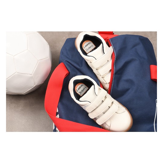 The image shows a white soccer ball, a pair of white athletic shoes with velcro straps, and a blue backpack with red details.