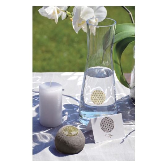 The image features a tranquil setting with a glass water pitcher, a white candle, a smooth stone, and a name card against a backdrop of a white tablecloth and green grass. Decorative symbols are placed on the stone and the card. White orchids are also visible, adding a serene touch to the scene.