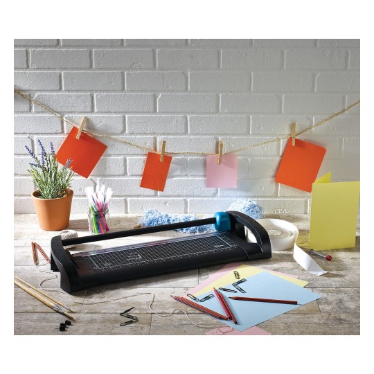 The image shows a crafting workspace with a paper cutter, colored sheets, scissors, paper clips, and a potted plant. The colored sheets are hung with clothespins on a string.