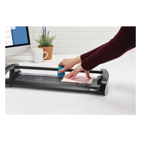 An image of a letter opener in use, while a person is cutting a sheet of pink paper. In the background, a computer and a potted plant can be seen.