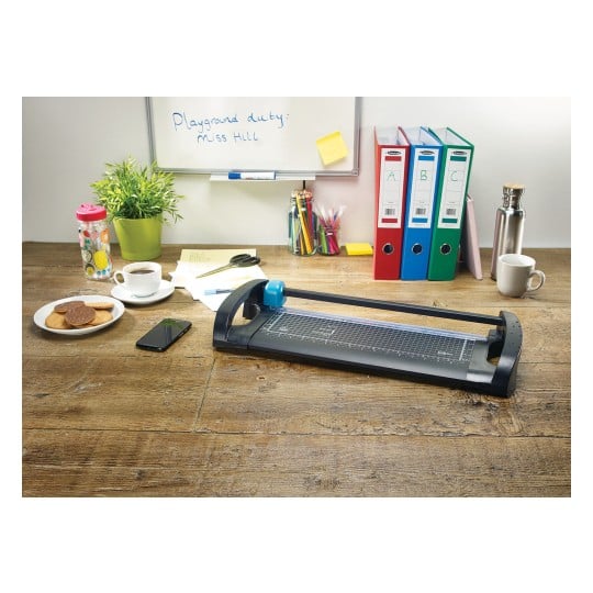 The image shows a workspace with a paper cutter, a cup of coffee, cookies on a plate, a smartphone, colored pens in a jar, folders, and a plant. There is also a whiteboard with a note.