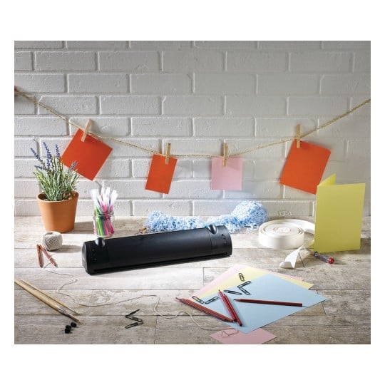 The image shows a creative workspace with various colored papers, pens, paper clips, a plant in a pot, and colored cards hanging on a string. There is also a device visible that is likely used for laminating.