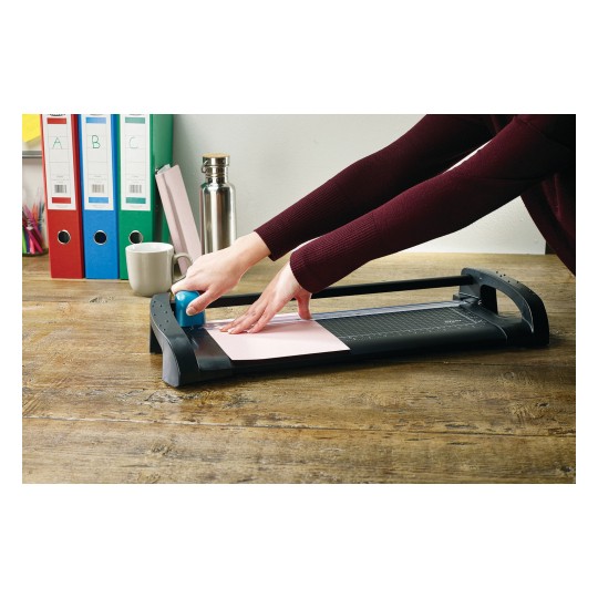 A hand cuts paper with a paper cutter on a wooden table. In the background, there are binders and a mug.
