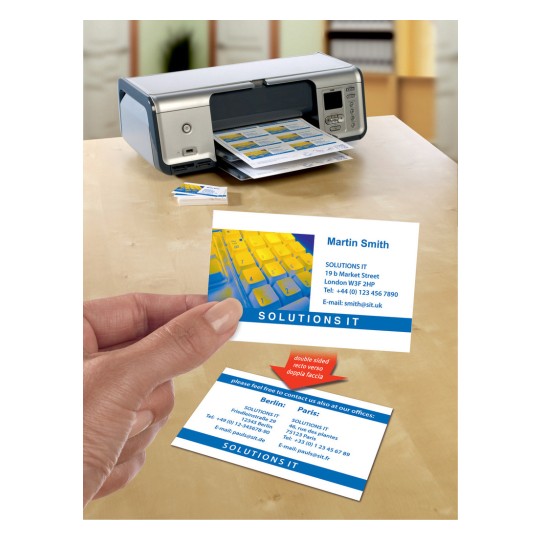 The image shows a printer with business cards printed next to it. A business card is in hand, highlighting contact information for 'Solutions IT' in London, Berlin, and Paris. A yellow keyboard is also visible.