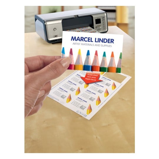 The image shows a printer and a hand holding a business card with the name 'Marcel Linder', while on the table there are labels with artistic styles. Colored pencils are a central element of the design.
