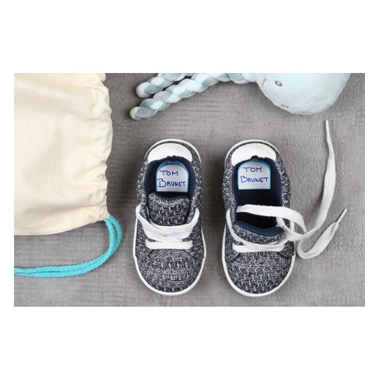 The image features a pair of small, gray knitted children's shoes with white laces, labeled 'TOM BRUNET.' Next to the shoes are a pastel-colored toy and a beige drawstring bag.