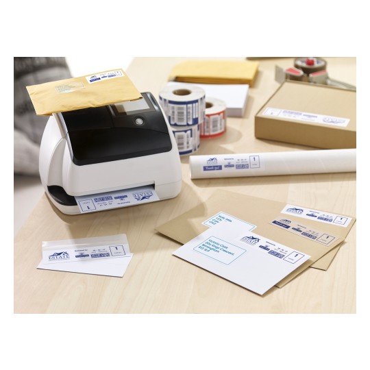 The image shows a label printer with a yellow envelope on top, surrounded by various envelopes, labels, a roll of tape, and a box on a wooden surface. The labels feature shipping information.
