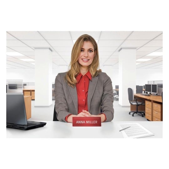 The image shows a woman dressed in a gray suit, sitting at an office desk. She has a nameplate with 'ANNA MILLER' in front of her and there is a laptop and some documents on the table. The background shows an office environment with office furniture.