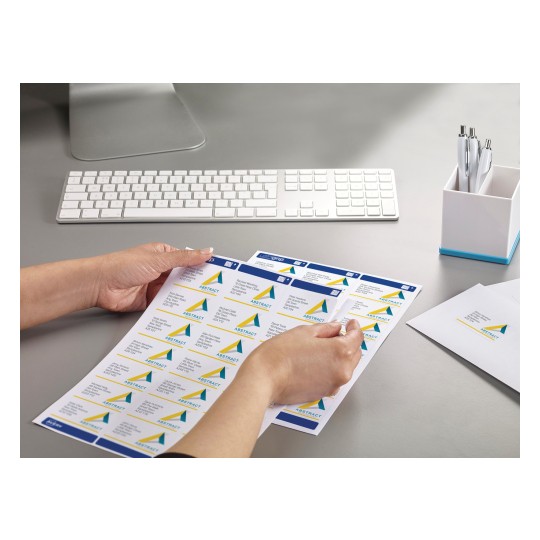The image features a person's hand holding a sheet of labels that include names and addresses, alongside a keyboard, a pen holder with pens, and some envelopes on a desk.