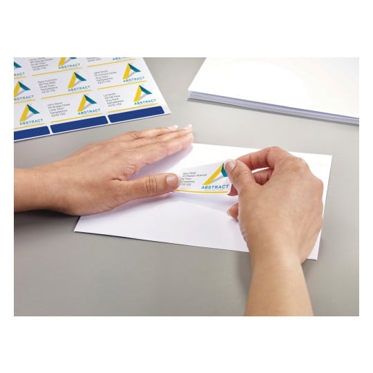 The image shows a person placing a label on an envelope. The label has a design with the word 'ABSTRACT' and includes a name and address. There are additional labels visible on a sheet, and blank envelopes are on the table.