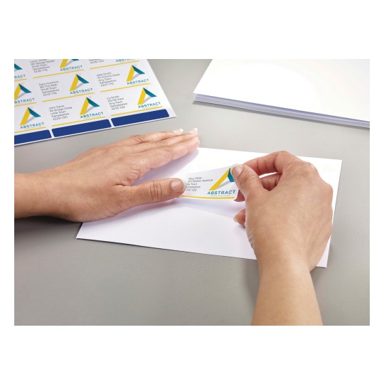 The image shows a hand applying a label to an envelope. The label has a logo and contact information from a company called 'ABSTRACT Architecture.' There are multiple labels on a sheet in the background, and some blank envelopes are also visible.
