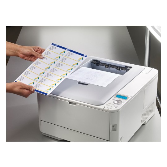 An individual is holding a sheet of printed labels in front of a laser printer. The printer is printing another document, and the labels feature a design with colored triangles and text.