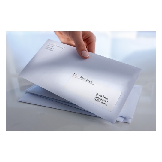 The image shows a hand holding a business envelope with a label. There are multiple envelopes stacked underneath. One envelope is addressed to Anna Berg in Berlin with a company name 'Mark Binder' visible.