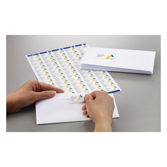 A person is applying a label from a sheet onto an envelope. The labels display a logo and text. Several labels are visible on the sheet, and there are more envelopes stacked nearby.