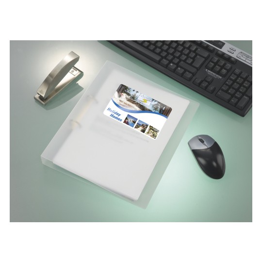The image shows a workspace with a transparent folder containing a brochure for holiday homes, a stapler, a computer keyboard, and a mouse on a sleek surface.