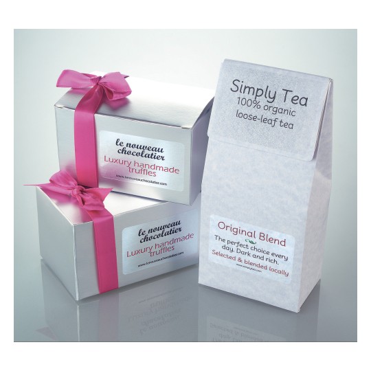 The image shows two gift boxes with luxury handmade truffles and a pack of loose tea. The boxes have a pink ribbon, while the tea organization is organic and locally selected.