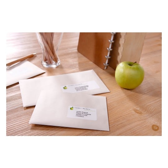 The image shows a wooden table with two addressed envelopes, a green apple, a glass container with wooden sticks, and a notebook. The envelopes have labels indicating names and addresses in France and Germany.