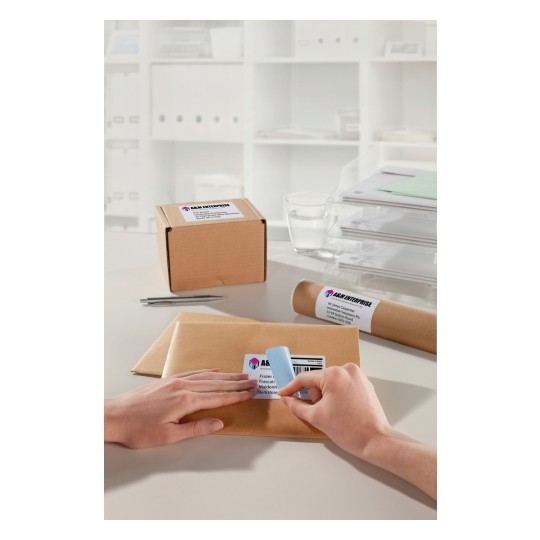 The image shows a desk with a few packaged shipments, including a box and an envelope. One hand is attaching a label to the envelope while another hand is involved. The background contains organized office supplies.