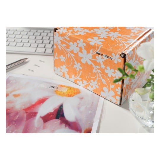 The image shows a colorful box with a floral pattern and a label with the name 'Anne Miller'. Next to it, there is a picture with the text 'prio A' and a computer keyboard in the background.