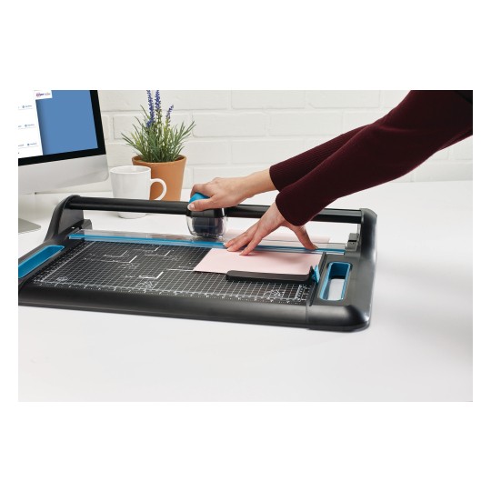In the image, a worktable with a cutting tool is observed. A person is using a cutter to work with a sheet of pink paper on the surface. There is also a computer and a potted plant in the background.