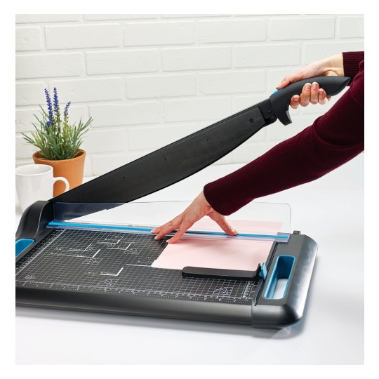 Image of a person using a paper cutter, placed on a table with a plant and a cup of coffee in the background.